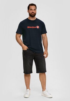 Marineblaue T-Shirt mit rotem "Sinalco"-Logo, kombiniert mit dunklem Denim-Shorts und weißen Sneakers. Baumwollmaterial mit einem einfachen Rundhalsausschnitt-Design.