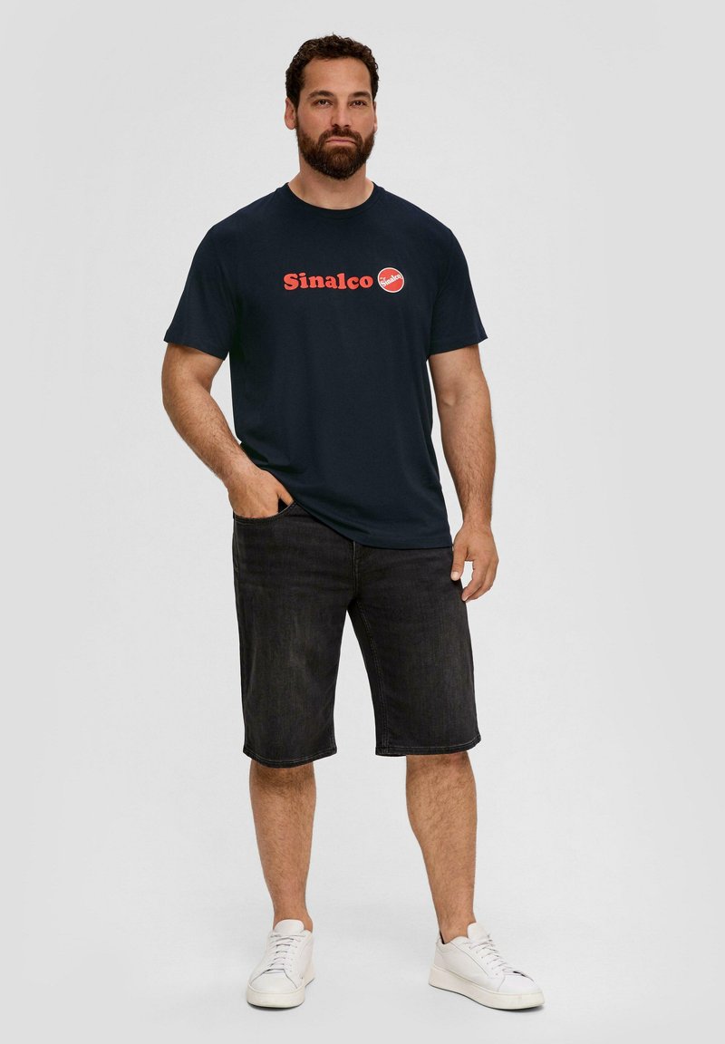 Marineblauwe t-shirt met rood "Sinalco" logo, gecombineerd met donker denim korte broek en witte sneakers. Katoenen materiaal met een basic ronde hals ontwerp.