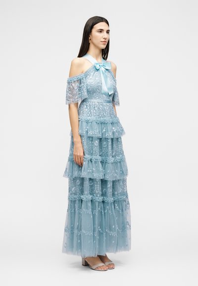 Γυναίκα με ανοιχτό μπλε φόρεμα από δαντέλα με στρώσεις και κεντήματα λουλουδιών, μανίκια off-shoulder και σατέν φιόγκο στο ντεκολτέ, όρθια πλάγια.