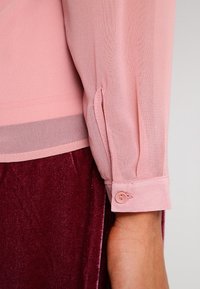 Ljusrosa blus med genomskinlig textur och knappade ärmar, tillsammans med en bourgogne-färgad sammetstyg nedtill, med en mjuk och slät yta.