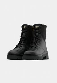 Para czarnych skórzanych botków na sznurowadła z grubymi gumowymi podeszwami i zamkami błyskawicznymi po bokach, przedstawionych na jednolitym jasnym tle.