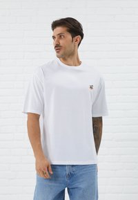 Weißes Baumwoll-T-Shirt mit kurzen Ärmeln, das ein buntes, gesticktes Design einer Palme und eines lächelnden Gesichts auf der linken Brust zeigt.