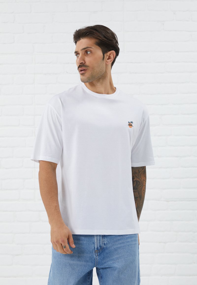 Weißes Baumwoll-T-Shirt mit kurzen Ärmeln, das ein buntes, gesticktes Design einer Palme und eines lächelnden Gesichts auf der linken Brust zeigt.