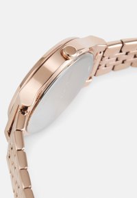 Roze gouden horloge met een gepolijste kast, ronde vorm en een gemofde metalen armband. Is voorzien van een zij kroon en een minimalistisch ontwerp.