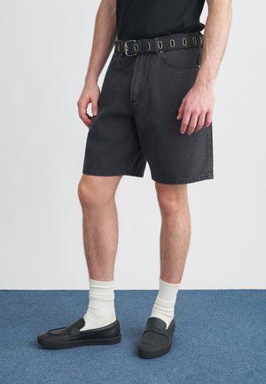 Μαύρα σορτς από τζιν με μπροστινή τσέπη, σε συνδυασμό με μια μαύρη ζώνη με κυκλικές τρύπες. Φοριούνται με άσπρες ριμπ κάλτσες και μαύρα παπούτσια χωρίς κορδόνια.