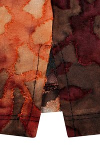 Strukturierter Stoff in warmen Tönen von Orange, Braun und Burgunder, mit fransigen Kanten und einem floralen Muster, das kontrastierende Nähte aufweist.