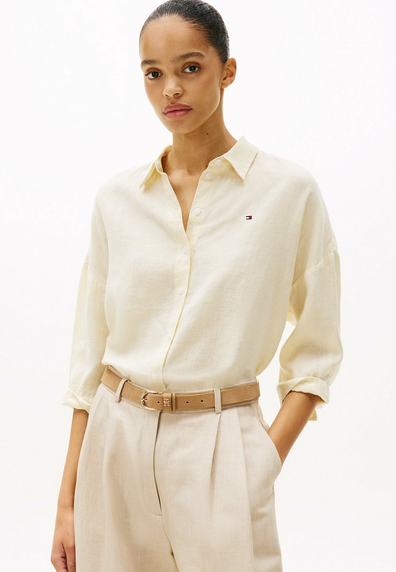 Camicia button-up gialla chiara con una vestibilità rilassata, dotata di un piccolo logo sul petto, abbinata a pantaloni beige e una cintura coordinata.