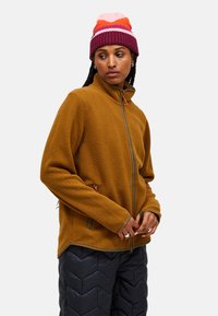 Giacca in pile marrone con tasche con zip e rifiniture in contrasto, abbinata a un berretto di maglia multicolore, indossata sopra pantaloni trapuntati neri.