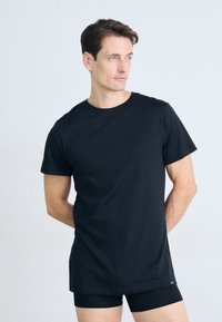 Man standing with hands behind back, wearing a plain black t-shirt and black shorts, looking slightly to the right against a white background.