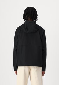 Schwarze wasserdichte Jacke mit Kapuze, entspannter Schnitt und nahtlosem Design; glatte Textur mit minimalistischem Detail auf der Rückseite.