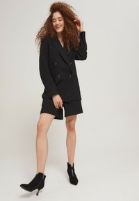 Blazer negro de doble botonadura combinado con pantalones cortos negros. El conjunto presenta un diseño elegante, una textura suave y una silueta ajustada. Botines negros hasta el tobillo completan el look.