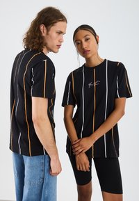 Svarta T-shirts med vertikala ränder i vitt och orange. Tyget är bomull med en avslappnad passform och har en liten logotyp på framsidan.