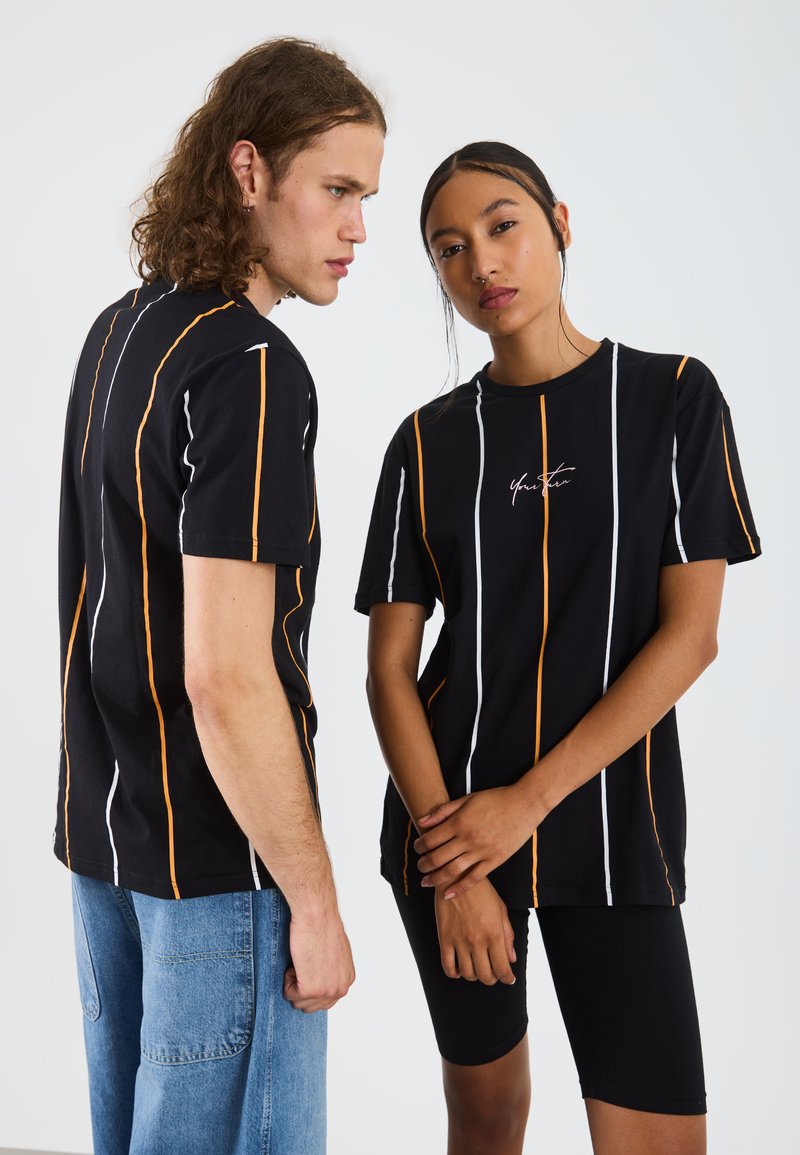 Svarta T-shirts med vertikala ränder i vitt och orange. Tyget är bomull med en avslappnad passform och har en liten logotyp på framsidan.