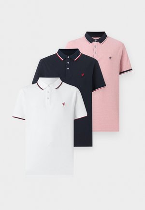 Άνδρας στέκεται σε λευκό τοίχο φορώντας λευκό πόλο με κόκκινο περίγραμμα και γκρι τζιν με μαύρα παπούτσια, ένα χέρι στην τσέπη.