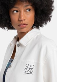 Veste en denim blanc avec un col classique, des boutons en argent et un motif brodé noir sur la poitrine gauche. Texture lisse.