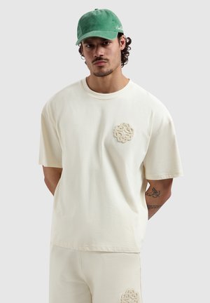 Mann trägt cremefarbenes oversized T-Shirt und Shorts mit strukturiertem Logo, grüne Kappe, steht mit den Händen hinter dem Rücken vor hellem Hintergrund.