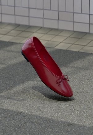 Jeden błyszczący czerwony baletowy balerinek z małą kokardką na strukturalnej szarej nawierzchni obok wyłożonej kafelkami ściany.