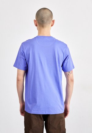 Osoba z ogoloną głową, ubrana w gładki fioletowy t-shirt i brązowe spodnie, widziana z tyłu na jednolitym białym tle.