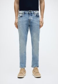 Φωτεινό μπλε τζιν παντελόνι με ίσια γραμμή, σχεδιασμό με πέντε τσέπες και διακριτική ξεθώριαση. Συνδυάζεται με μπεζ αθλητικά παπούτσια.