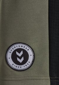 Olivgrön sportklädesväv med svart texturerad panel, med en rund etikett med svart och vit design som säger "SPORTKLÄDER SEDAN 1923."