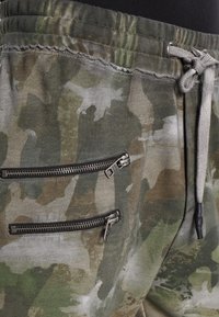 Tarnmuster-Jogging-Shorts aus grünem und grauem Stoff mit einem Kordelzugbund, ausgestattet mit zwei Reißverschlusstaschen mit silberner Hardware.