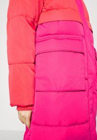 Veste rembourrée rose avec une texture lisse, présentant une section supérieure corail contrastante, des poches latérales et des poignets élastiques. Design en blocs de couleurs vives.