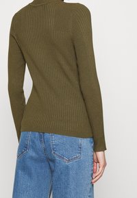 Olivengrüner gerippter Pullover mit taillierter Form und langen Ärmeln, kombiniert mit hellblauen Jeans aus Denim mit klassischen Gesäßtaschen.