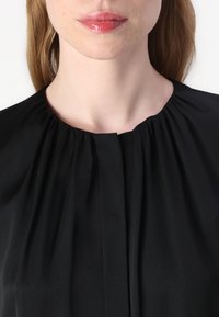 Blusa nera con dettaglio di collo arricciato, caratterizzata da pieghe e un tessuto liscio e testurizzato. Design semplice e pulito con colletto rotondo.