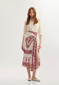 Chemise crème à boutons accompagnée d'une longue jupe portefeuille rouge à motifs complexes et fendillée jusqu'à la cuisse. Ballerines plates beiges.