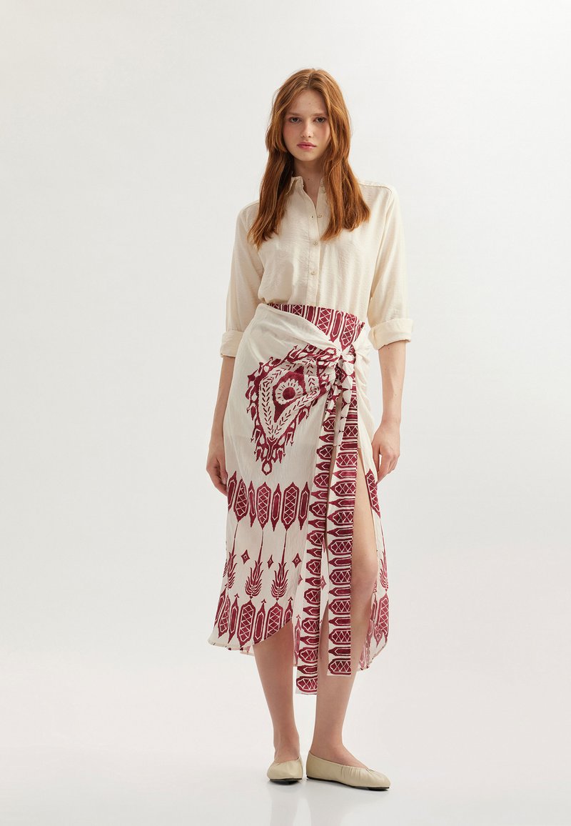 Chemise crème à boutons accompagnée d'une longue jupe portefeuille rouge à motifs complexes et fendillée jusqu'à la cuisse. Ballerines plates beiges.