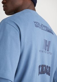 Svijetloplavi pamučni majica s printanim logotipima na leđima: "HOLZWEILER SKICLUB," "WINTER SPOT," i stiliziranim "BDG." Mekana tekstura.