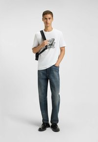 Witte kortslung t-shirt met een zwart grafisch ontwerp, gecombineerd met donkerblauwe spijkerbroek en zwarte schoenen, met een zwarte schoudertas.