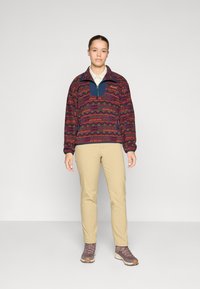 Pullover in fleece multicolore con motivi geometrici, colletto a mezza zip e dettagli blu navy uniforme, abbinato a pantaloni beige e scarpe da trekking marroni.
