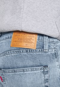 Ljusblå denimjeans med en läderetikett på bakfickan, med präglad text och en design som föreställer djur och verktyg.