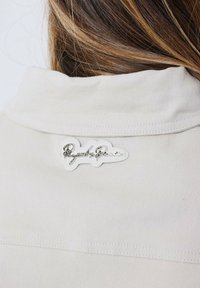 Crème-kleurige stoffen kraag met een metalen, cursieve logo-tag. De kraag is glad met een nette stikselafwerking.