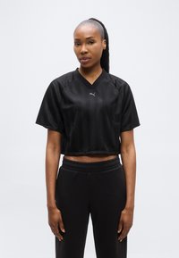 Γυναίκα με σκούρα μαλλιά σε αλογοουρά, φορόντας μαύρο κοντό αθλητικό μπλουζάκι με το λογότυπο της Puma και μαύρο παντελόνι, στέκεται μπροστά σε ένα απλό φόντο.