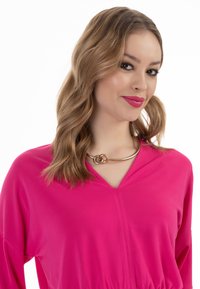 Blusa rosa a maniche lunghe con scollo a V, realizzata in tessuto texturizzato e con un collier a cerchi intrecciati dorati.