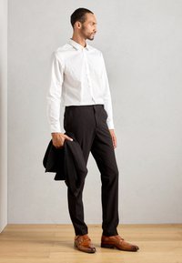 Hvit skjorte med knapper, svarte bukser, brune loafers. Modellen holder en svart blazer. Glatte teksturer, skreddersydd passform og rene linjer.