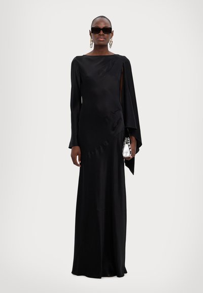 Vestido negro de manga larga con un diseño tipo capa fluido, de tela suave y con una abertura lateral. Combinado con gafas de sol de gran tamaño y un bolso metálico.