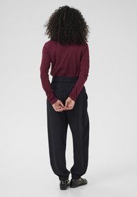 Haut maroon à manches longues associé à un pantalon noir ample, avec taille à cordon de serrage et poignets élastiques. Des chaussures noires complètent l'ensemble.