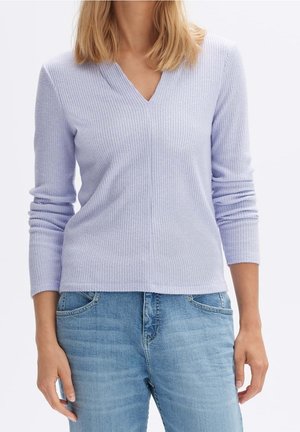 Vrouw draagt een lichtpaarse geribbelde lange mouw V-hals trui gecombineerd met lichtblauwe high-waisted jeans, staand met ontspannen handen.