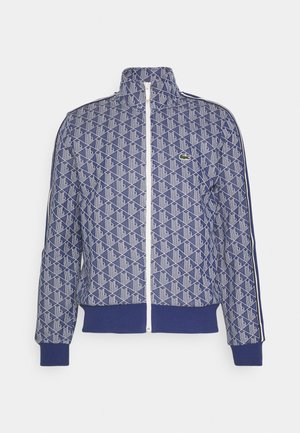 Jūras spēku sporta žakete ar ģeometrisku rakstu baltā krāsā. Iespējas augsts apkakles, pilns rāvējslēdzis un ribotas aproces. Ietver logotipu kreisajā krūtī.