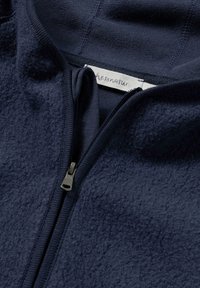 Marineblaue Fleece-Zip-Jacke mit weicher Textur, ausgestattet mit einem kontrastierenden Innenfutter und einem Markenlabel. Der Reißverschluss ist dunkel und funktional.