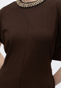 Haut marron à manches courtes avec une couture centrale et un col décoratif orné d'embellissements dorés et argentés sur une bordure noire.