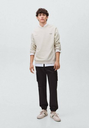 Beige sweatshirt met een ronde hals, gelaagd over een wit overhemd met knopen; gecombineerd met zwarte cargo-broeken en lichte sneakers.