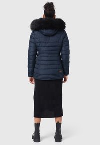 Marine pufferjack met een afneembare faux fur capuchon. Kenmerken zijn de geborduurde textuur, een ceintuur in de taille en een embleemdetail op de mouw. Zwarte geribbelde rok en chunky laarzen.