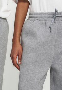 Άτομο με σκουρόχρωμο δέρμα που φοράει ανοιχτό γκρι φόρμα με ελαστική μέση και κορδόνι, ορατό χέρι που ακουμπά στο πλάι.