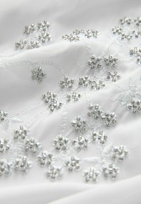 Tissu blanc avec des grappes complexes de perles argentées disposées en motifs floraux avec une broderie délicate en fil argenté.