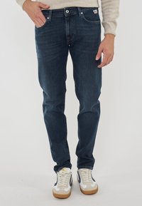 Uomo che indossa jeans blu scuro, maglione beige e sneakers bianche con dettagli beige e navy, in piedi di fronte a uno sfondo chiaro e uniforme.