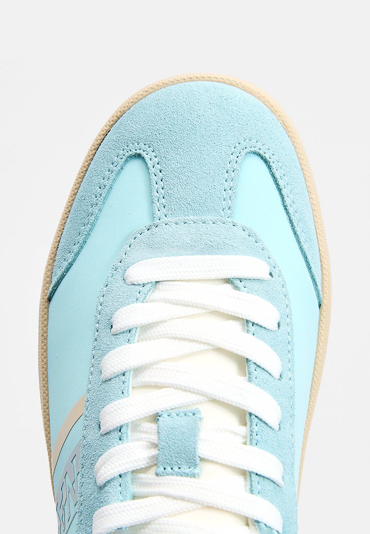 Zapatilla azul clara que combina materiales de ante y lisos, con cordones blancos y suela de goma beige. Punta redondeada con detalles destacados.